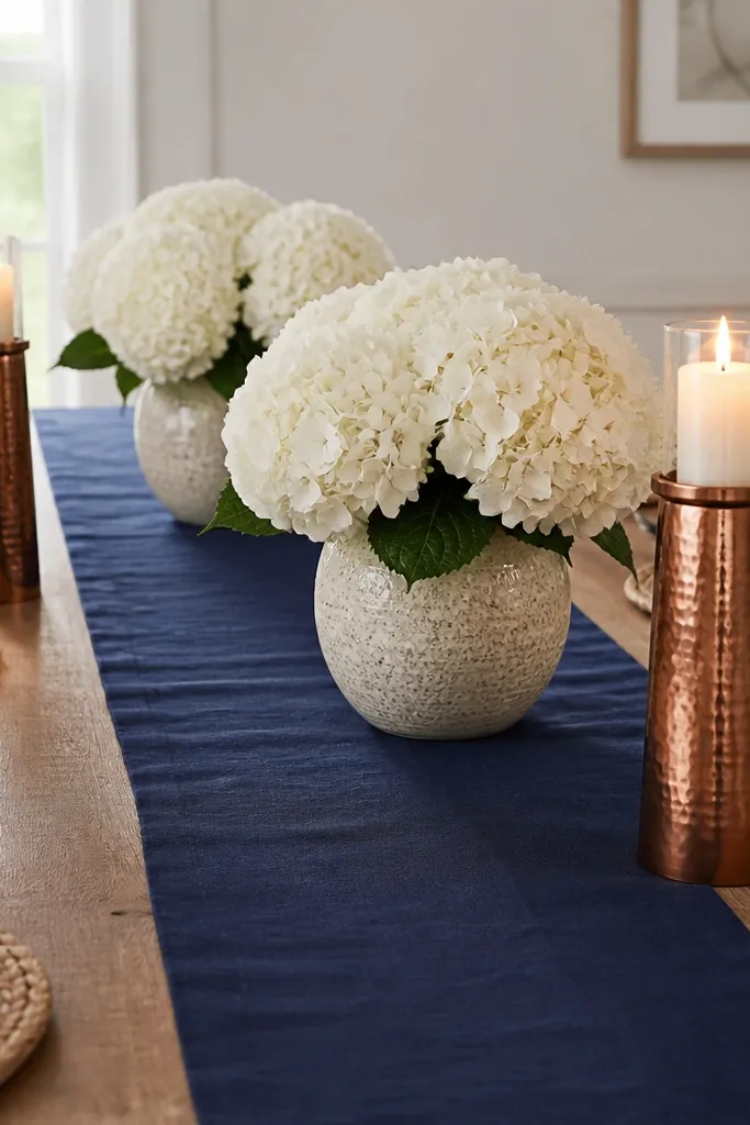 A navy runner runs down the table center. Two large white hydrangea-style faux clusters sit in matching vases. Copper candle holders flank the arrangement.