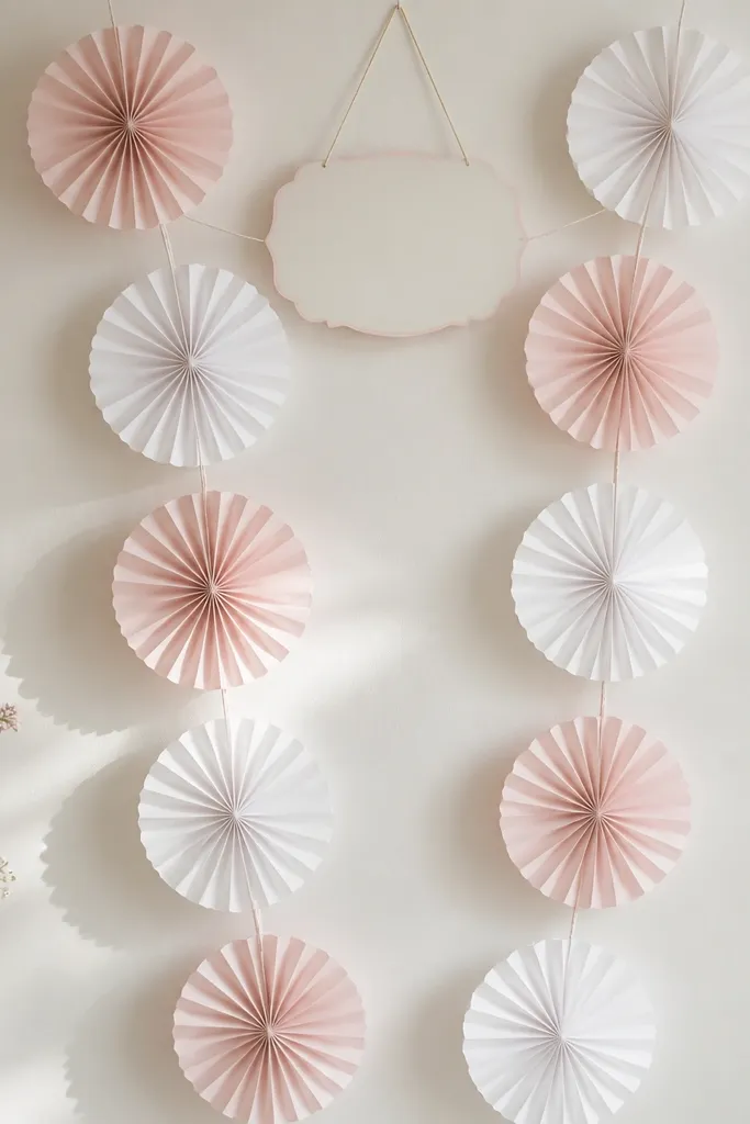 A vertical strip of alternating white and blush paper fans, each fan about 12 inches tall. The fans attach to thin string across a wall, with a small "Bride to be" sign centered near the top.