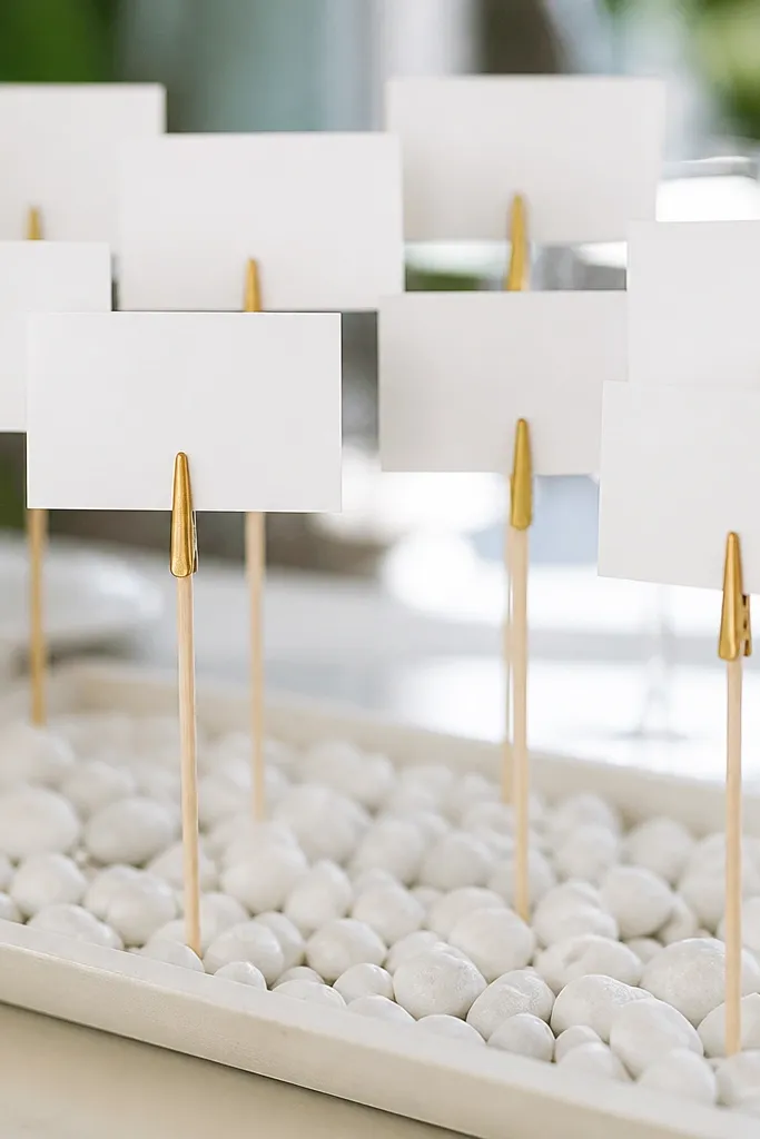 Thin wooden dowels with gold paint pen tips holding small place cards; the holders sit in a shallow tray with white pebbles.
