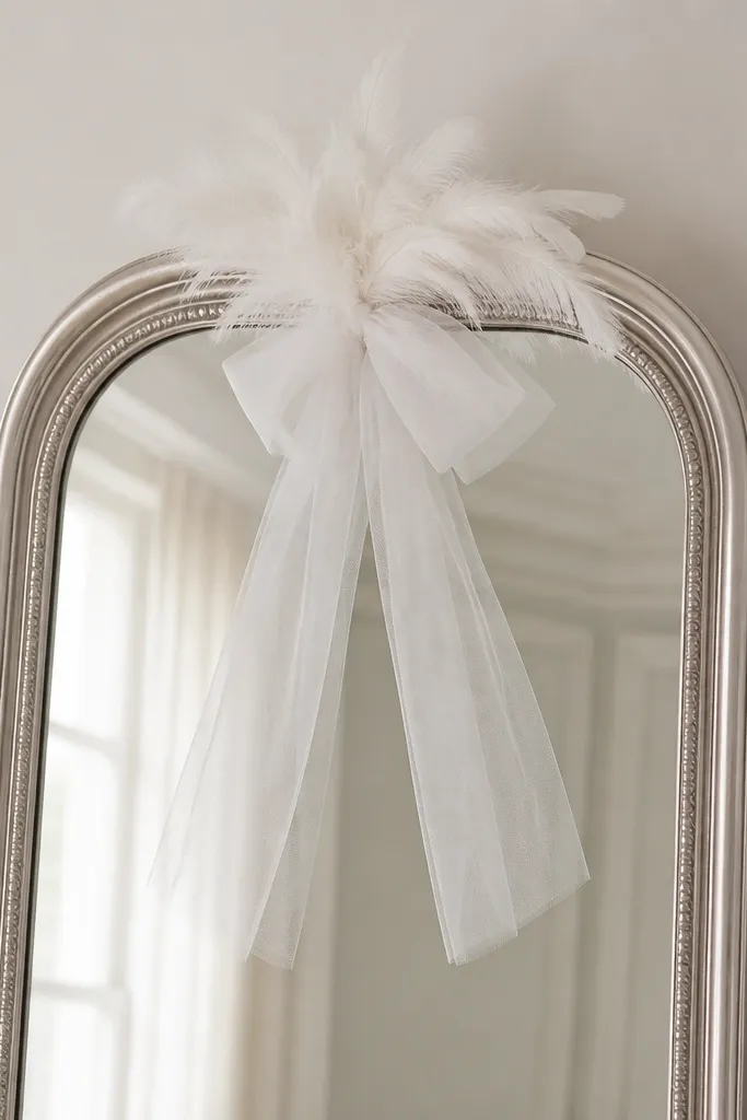 A silver standing mirror. A small cluster of white feathers sits at the top center. A soft white tulle ribbon loop hangs below the feather cluster, with two tails draping down.