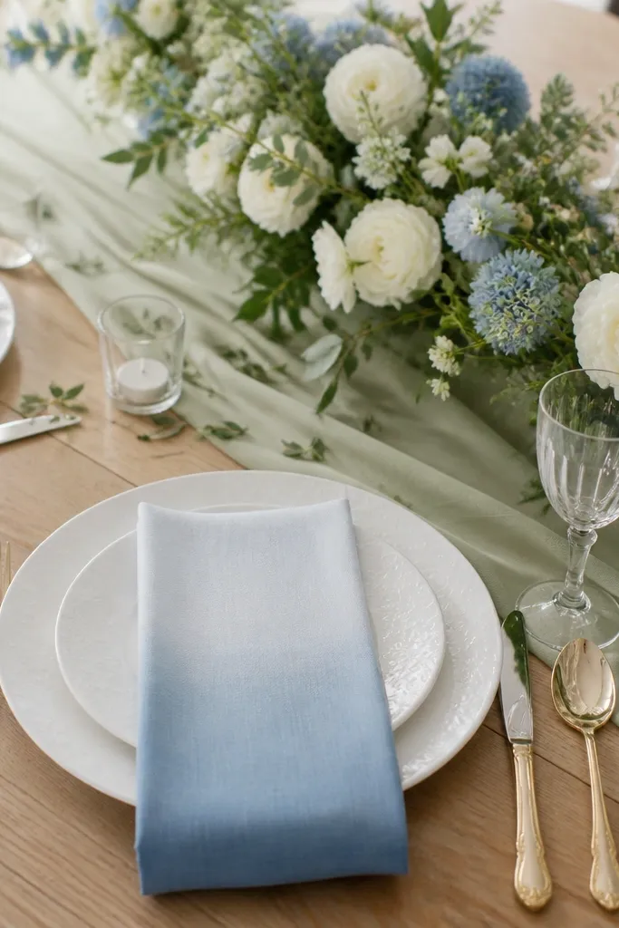 White plates with a faint floral pattern. Napkins show a dusty blue ombre effect from pale top to deeper bottom. A sage runner sits under a low arrangement of white and dusty blue flowers, with small green leaves scattered.