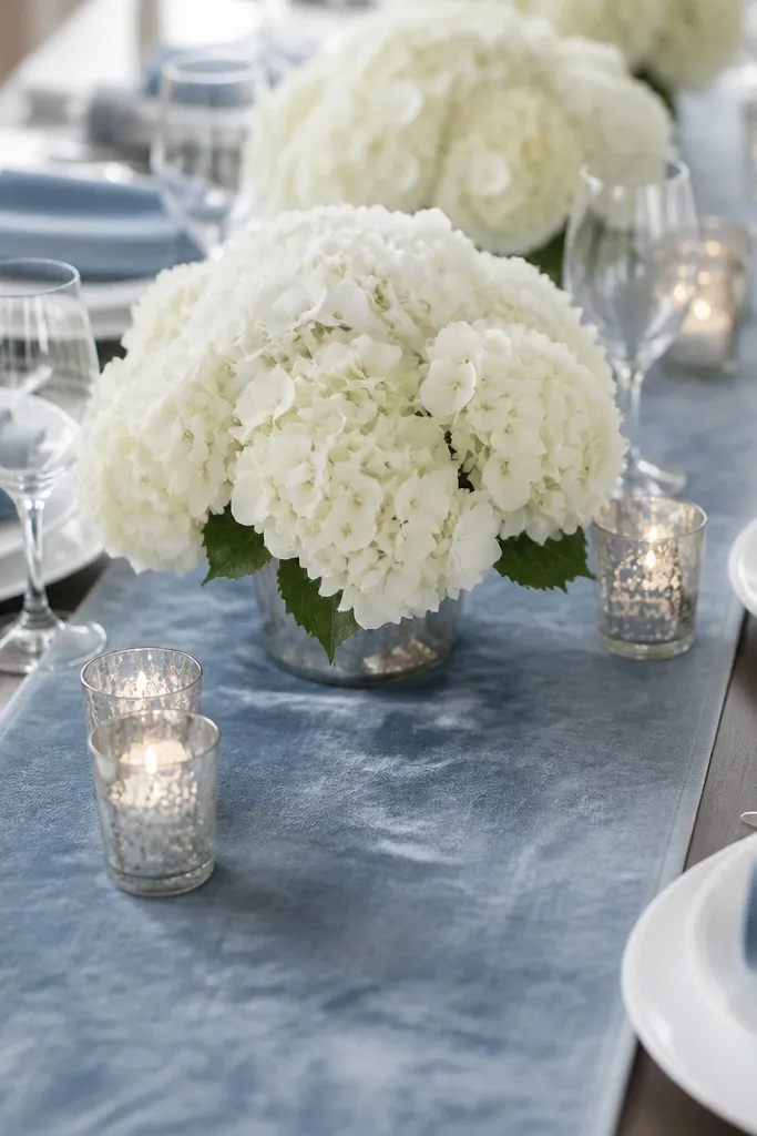 A dusty blue velvet runner down the center with soft texture. White hydrangea blooms sit in a low arrangement. Mercury glass votives reflect light, and place settings have white plates and dusty blue napkins folded into a soft roll.