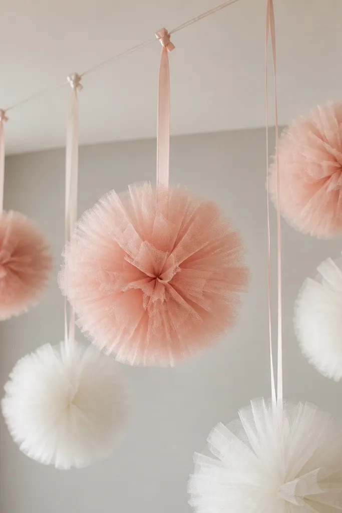 Several tulle pom-poms in blush and white hanging from a ceiling line, each pom attached to a ribbon string with a small knot.