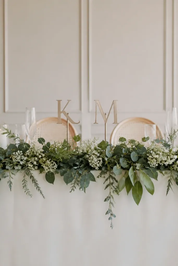 A sweetheart table with greenery garland across the front. Two monogram initials sit on stands near the center, with micro blooms tucked into the garland.
