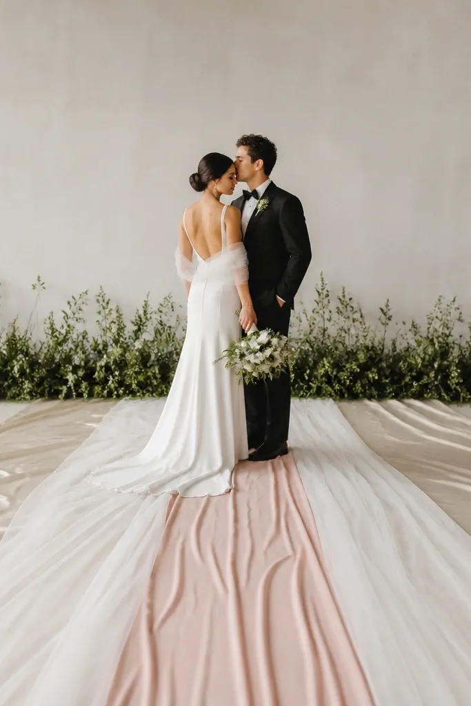 A long layered fabric setup across the stage floor: a champagne satin base, an ivory tulle layer above it, and a narrow blush crepe strip running down the center. Behind it, a simple greenery line traces the backdrop edge. The couple stands on the layered center strip.