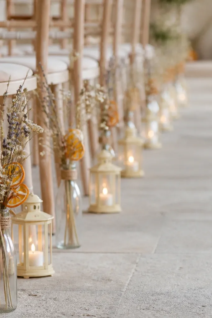 A ceremony aisle lined with slim glass bottles holding small dried stems and a few orange zest curls tied with twine, plus small cream lanterns between vases.