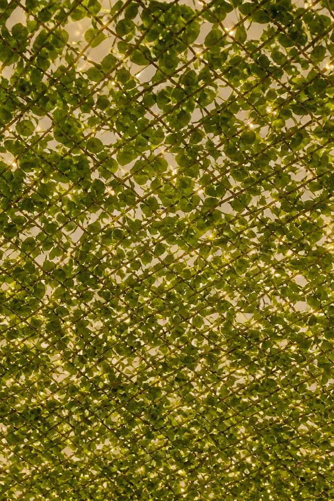 An overhead canopy made from green artificial vines and leaf garlands. Warm white lights are woven through the vine netting, creating a soft glow across the ceiling area. The scene looks full overhead with no dangling cords.