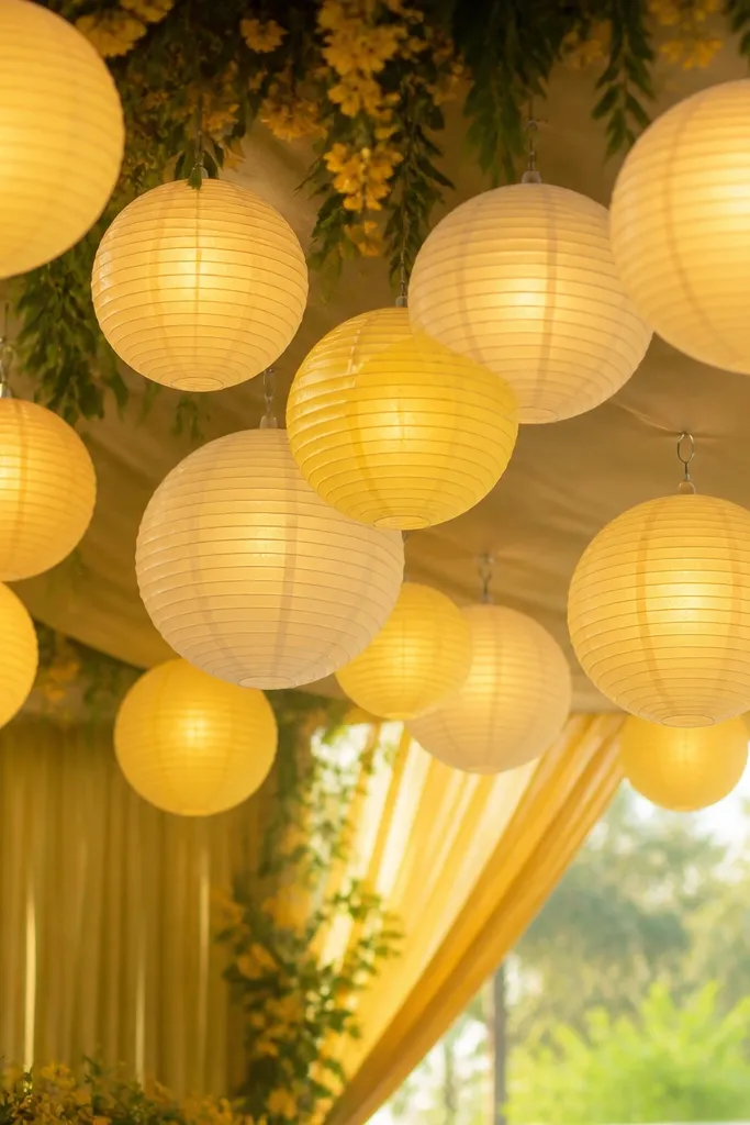 Hanging paper lanterns in cream and pale yellow colors. Warm micro fairy lights are placed inside each lantern, creating an even glowing interior. The lanterns are hung above the haldi area with visible hooks but no messy wires.