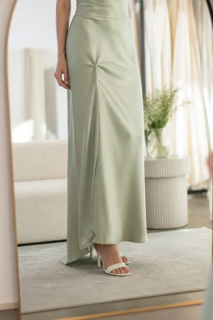 A fitting mirror shot with a sage gown hem pinned at ankle height while the model wears the wedding shoes; the hem falls cleanly with minimal extra fabric on the floor.
