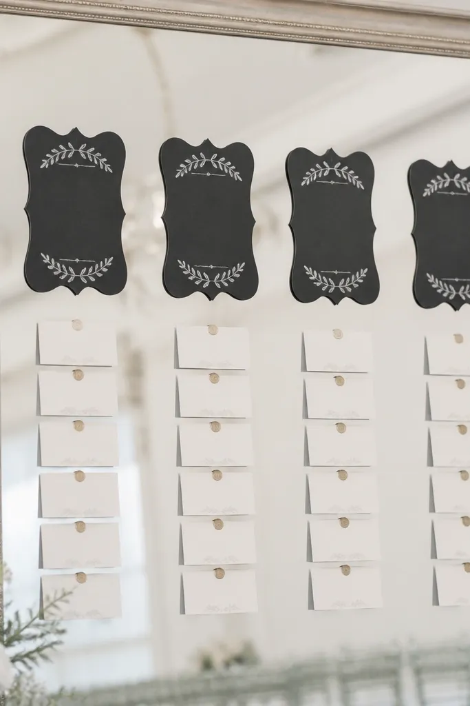 A silver framed mirror with matte chalkboard-style table headers. The headers look like chalk writing but are printed in a matte finish. Name cards sit neatly below, and the mirror reflection shows a soft, clean background with no sticky residue.