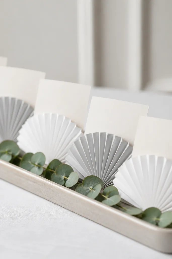 A row of small folded paper fans in soft white and pale gray, each fan holding a place card; the fans sit upright on a narrow tray with a single sprig of eucalyptus at the base.