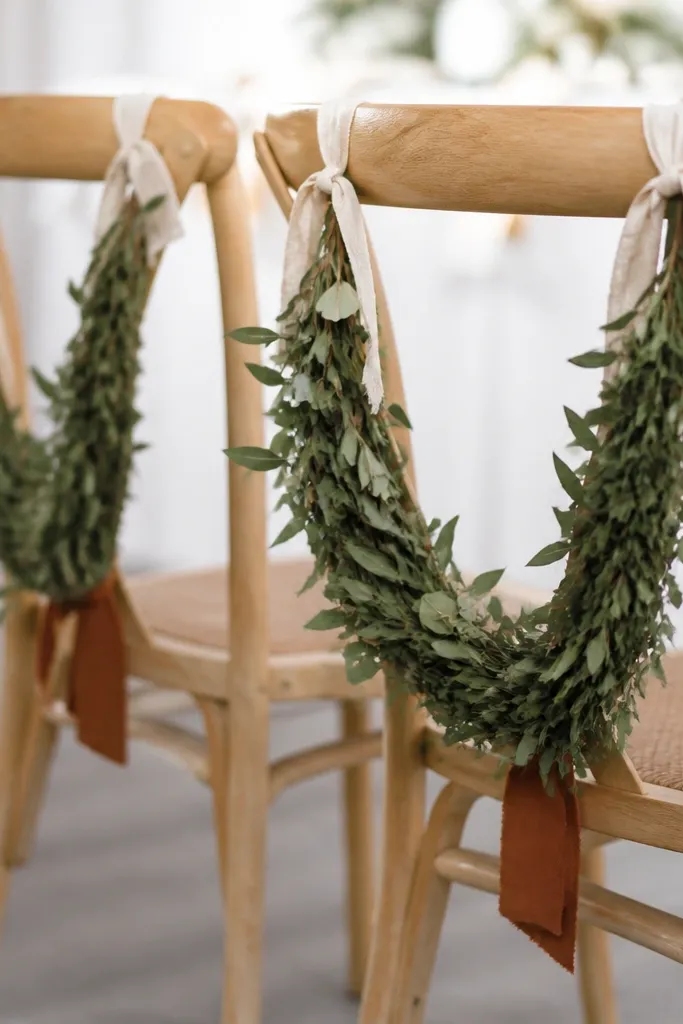 Two curved greenery pieces attached to chair backs using cream linen ribbon, with small rust tags tied near the bottom.