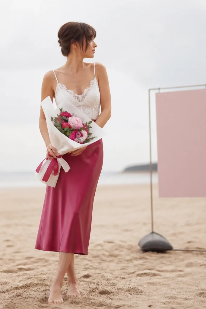 A woman wears a raspberry satin slip skirt that falls mid-calf and a white lace camisole. She holds a bouquet wrapped in white paper with a raspberry ribbon and stands on sand with a small weighted sign placed behind her.