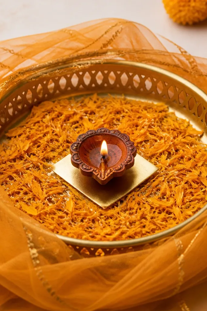 A tray with saffron net fabric draped loosely over the outer rim like a soft curtain, a marigold petal scatter in the center, and a diya placed in the middle with a small gold paper strip.