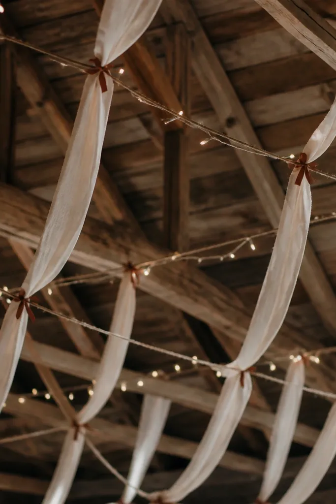 Thin jute ropes and cream linen strips hang from a barn ceiling in a loose grid. Small rust-colored ribbon ties connect some intersections. The strips sway slightly, with fairy lights tucked along the ropes.
