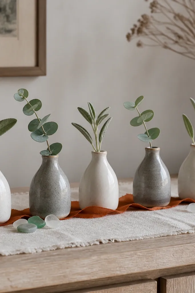 A lineup of small stoneware bud vases in off-white and gray on a sideboard. Each vase holds a single sage sprig or eucalyptus leaf. The vases sit on a linen runner with a few scattered sea-glass colored stones and a rust ribbon across the center.