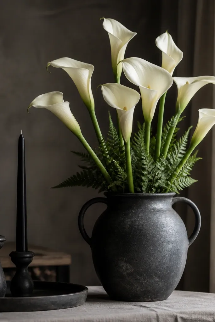A charcoal stoneware jug with a wide mouth sits on a table. Inside are cream calla lilies with long stems and a few fern fronds at the base. The jug sits near black taper candles on a matching stoneware plate.