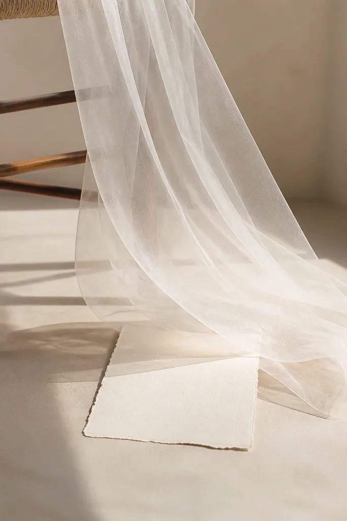 A sheer white veil fabric hangs from a chair; the save the date card is placed underneath, and the fabric lifts slightly as if floating, with soft daylight.