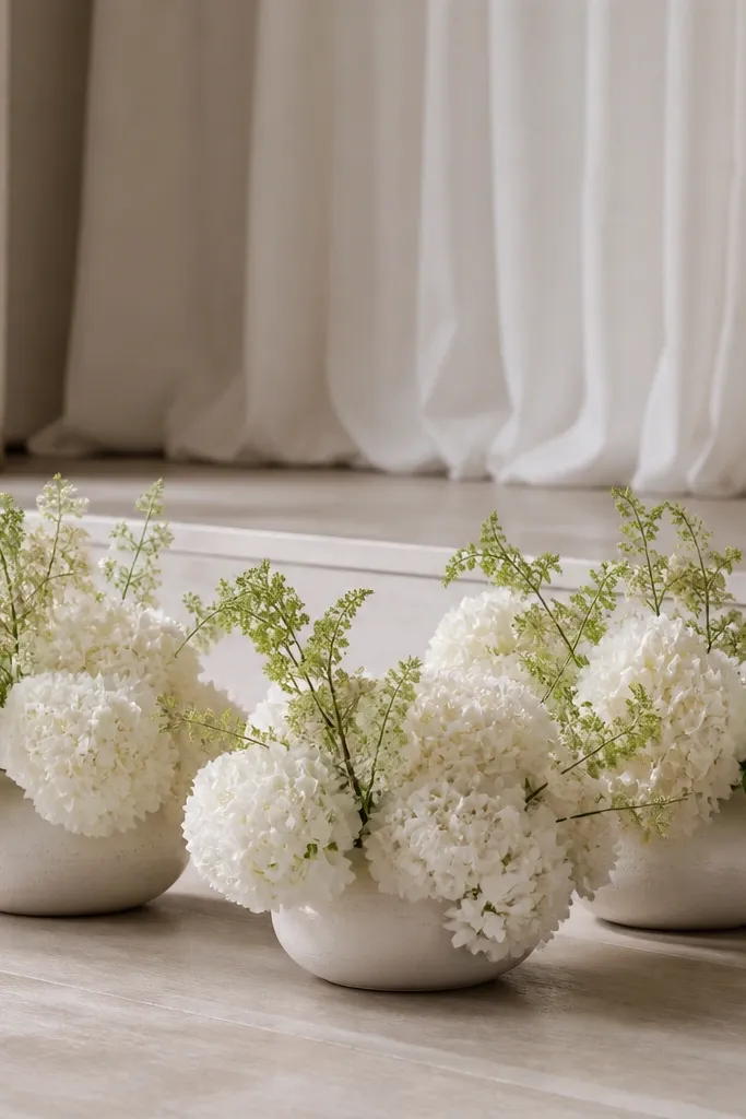 Three low ceramic vases on the stage floor filled with white hydrangea-style artificial sprays and a few pale green stems.