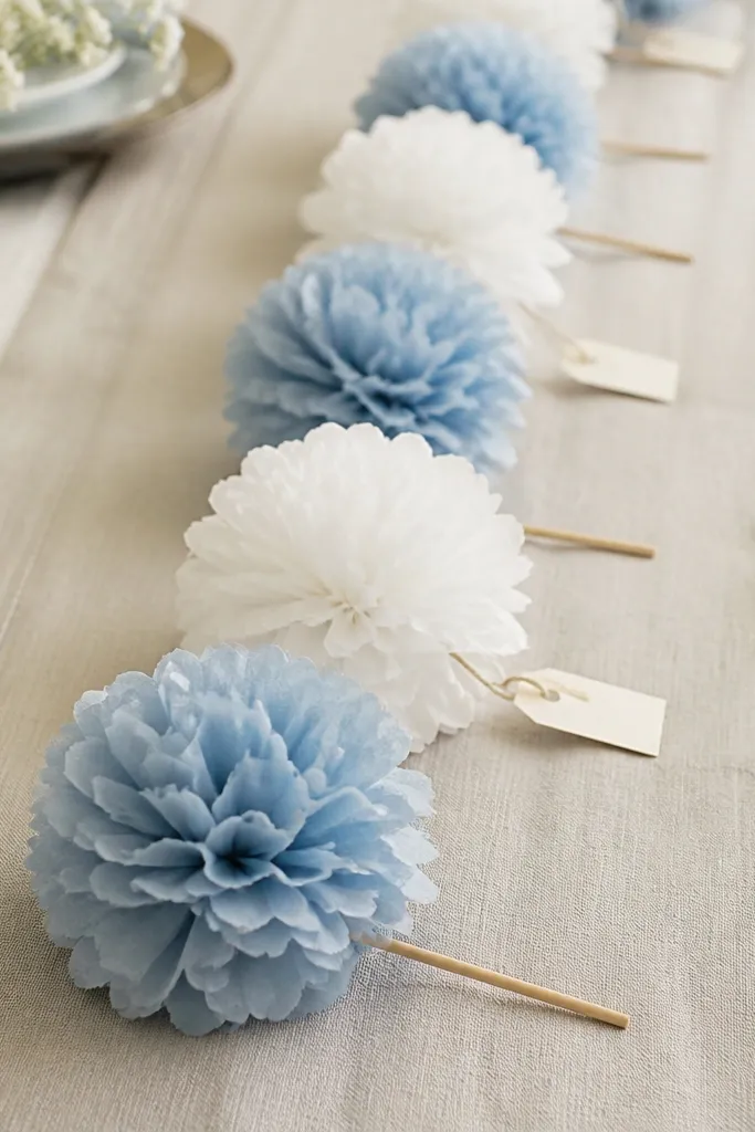 Tissue paper pom pom bows in white and dusty blue attached to small wooden skewers, arranged in a line along a runner, with a handwritten tag on each.