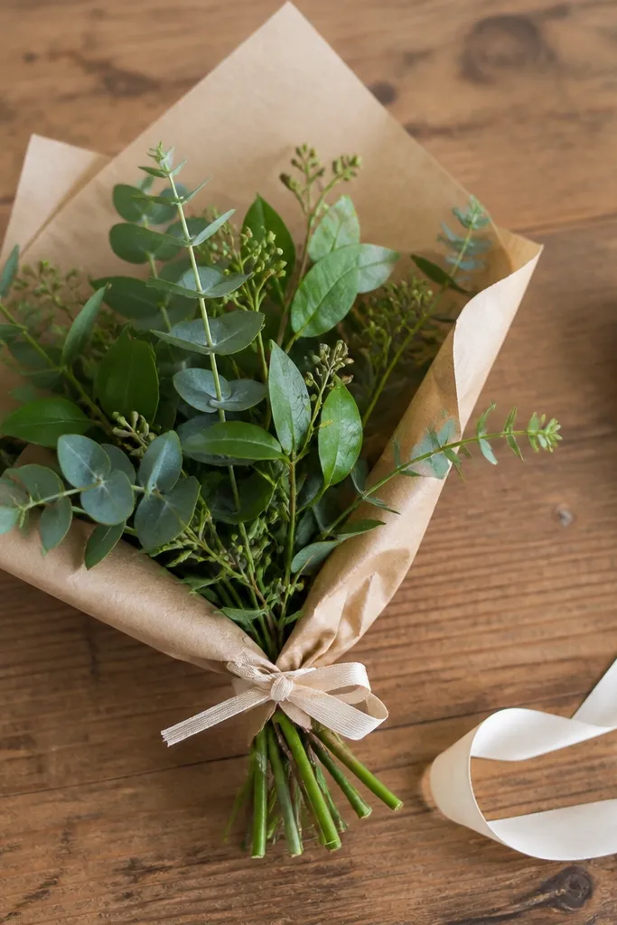 A small hand-tied bouquet with mixed greens (eucalyptus-like leaves, ruscus, and small sprigs) wrapped in brown kraft paper and tied with a thin ribbon. The bouquet sits on a wooden table with a cream ribbon nearby.