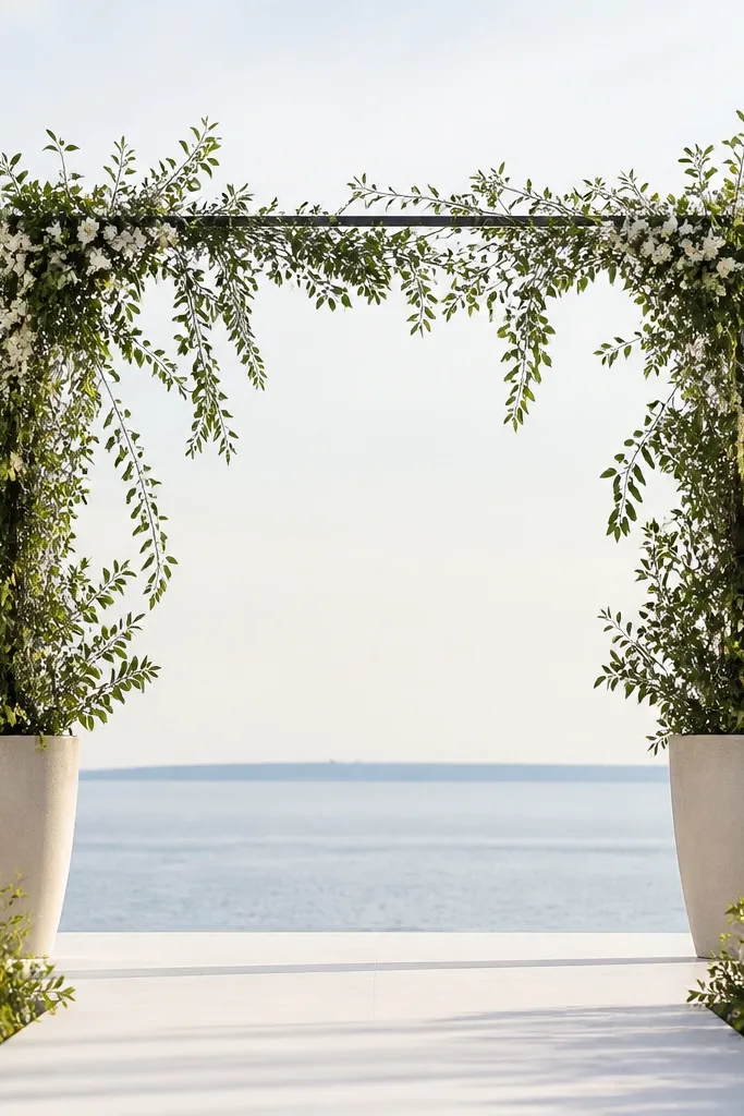 A ceremony backdrop made from two tall potted plants on either side of a simple frame, with trailing greenery draped between them. There are small white blossoms scattered near the top, and the ground has a clean walkway.