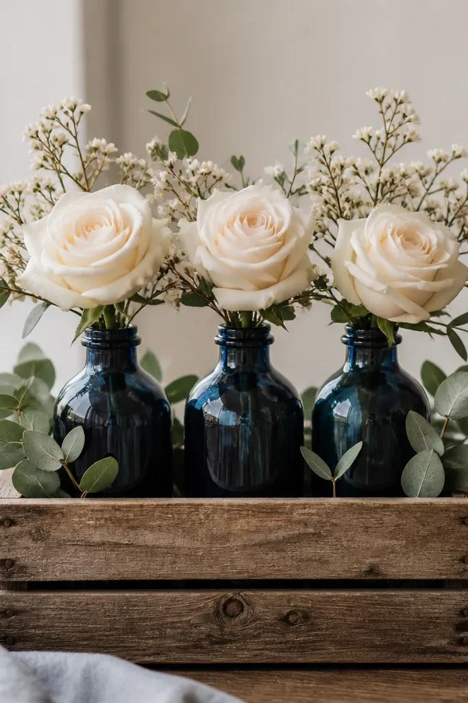 A small reclaimed wood crate on a table, holding three navy glass bottles. Cream roses and small sprigs spill gently from the bottle openings, with green leaves filling the crate corners.