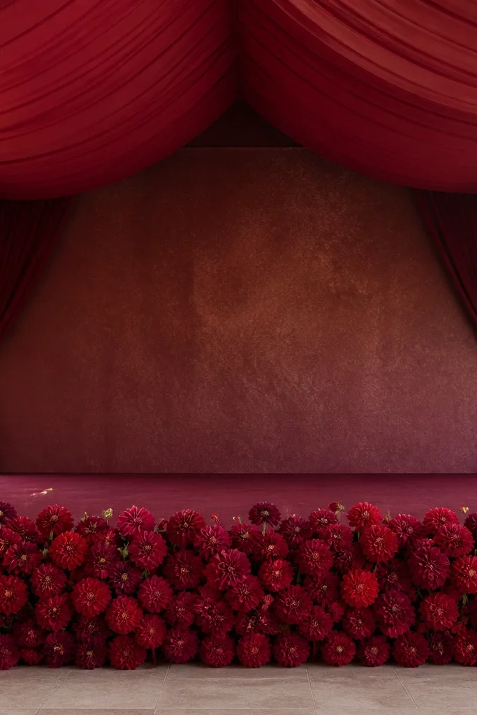 A stage front border made of dense red dahlia pom-pom clusters, with a canopy of deep red and wine fabric stretched above; the center backdrop is a simpler textured panel.