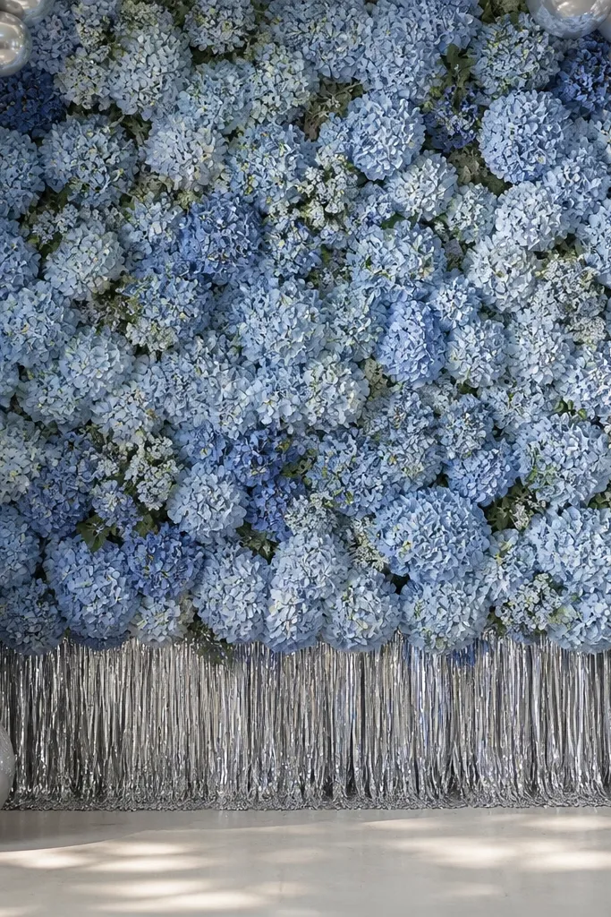 A blue floral backdrop using hydrangea-like clusters, with silver balloon clusters at the corners and a thin silver fringe curtain along the lower edge.