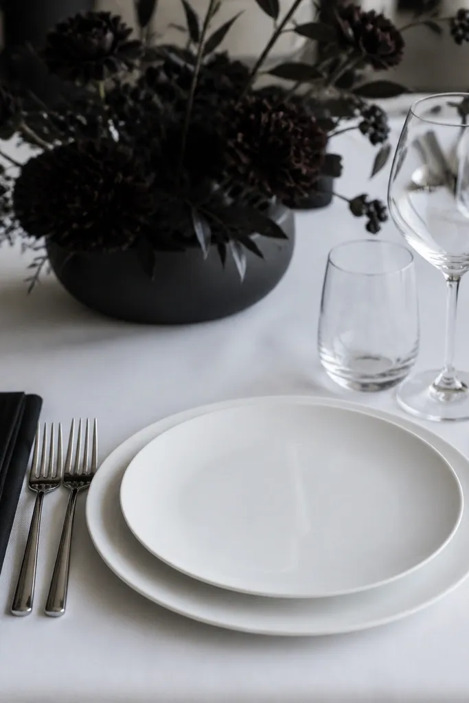 A monochrome place setting: white dinner plate on a white charger, a folded black napkin placed neatly to the left, polished silver cutlery, and clear water and wine glasses. The centerpiece is dark and minimal, keeping the focus on the plates.