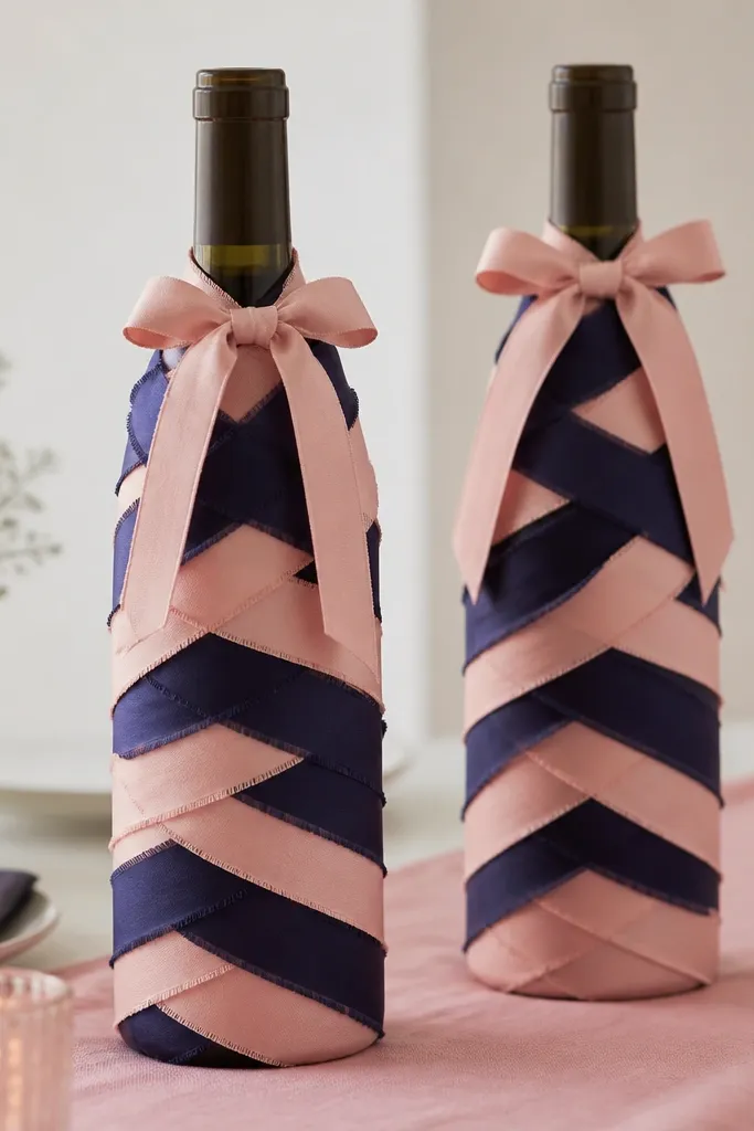 Wine bottles wrapped with layered fabric strips that look like overlapping ribbon. Colors alternate between navy and blush pink. The strips are tied at the top with a small bow, and the bottles sit on a pink table runner.
